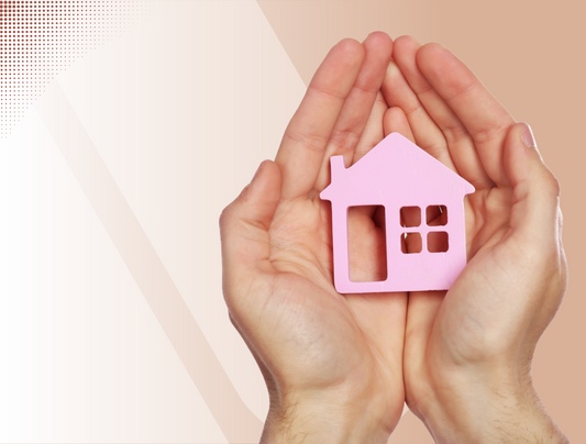 Zwei Hände sind aneinandergelegt und halten einen kleinen, rosafarbenen, hausförmigen Ausschnitt mit Fenstern und Türen vor einem Hintergrund mit hellem Beige und weißem Farbverlauf.