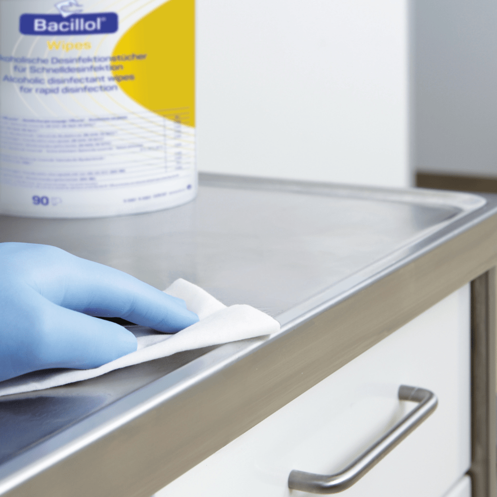 Une main gantée nettoie une surface métallique à l'aide de lingettes désinfectantes « Hartmann Bacillol® Wipes » de Paul Hartmann AG, en sachet à fond plat, 90 lingettes | pièce (1 pièce). Le contenant renfermant les lingettes Bacillol est partiellement visible à l'arrière-plan et présente quelques éléments de texte et de design sur l'étiquette. La surface métallique semble faire partie d’un meuble muni d’un tiroir.