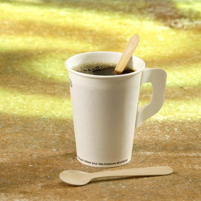 Un gobelet en carton blanc avec une anse contient du café noir et une cuillère à café en bois PAPSTAR "pure", 11 cm de PAPSTAR GmbH comme agitateur ; à côté se trouve une autre cuillère en bois sur un fond jaunâtre flou.