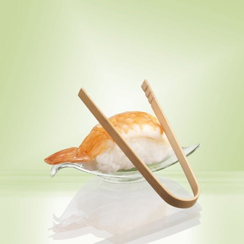 Une paire de 10 cm PAPSTAR Pinces à amuse-bouches en bambou de PAPSTAR La GmbH repose sur un plat en verre à côté de nigiris aux crevettes – idéal pour servir des en-cas ou des amuse-bouches lors d'une fête – sur un fond présentant un dégradé de vert pâle.
