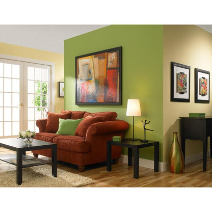 Un salon avec des murs jaunes et verts peints avec le ruban de masquage ScotchBlue™ "Multi-Surface" de 3M Deutschland GmbH, un canapé rouge avec des coussins verts, une table basse noire, une table d'appoint avec lampe, de l'art abstrait, un sol en bois et des portes vitrées.
