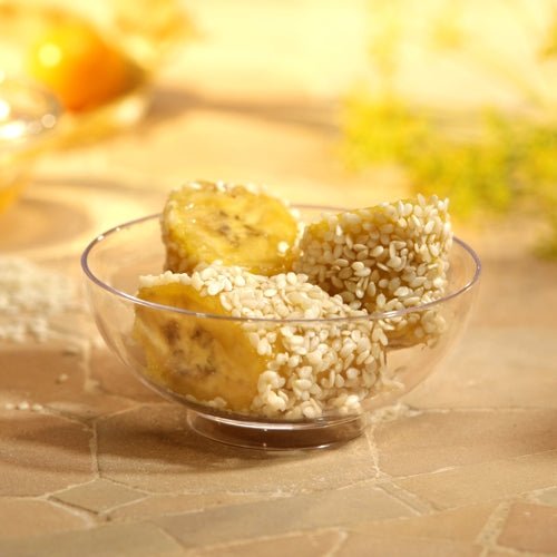 Trois morceaux de banane avec du sésame blanc sont servis dans un bol à fingerfood Starpak 50 (PS transparent), placés sur une surface carrelée claire avec un arrière-plan légèrement flou - idéal pour les événements.