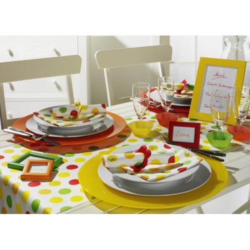 Une table pour deux personnes avec des sets de table jaunes et orange, des assiettes blanches, PAPSTAR 20 serviettes 33x33 cm de PAPSTAR GmbH, des verres, des cartes de table encadrées, une nappe à pois rouge-jaune-vert et une décoration ainsi qu'un menu encadré en arrière-plan.