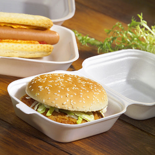 Un hamburger dans un petit pain au sésame avec de la salade et un steak, servi dans une boîte à hamburger Starpak 100 avec couvercle rabattable, XPS, 7,5 x 12 x 12 cm blanc - idéal pour le catering, avec des hot-dogs à proximité et des légumes-feuilles sur une surface en bois.