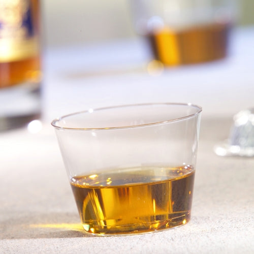 Un gobelet Starpak de 15 pièces, PS 0,1 l parfaitement clair, contient une petite quantité de liquide ambré sur une surface claire, tandis qu'en arrière-plan un autre gobelet et une bouteille sont flous.