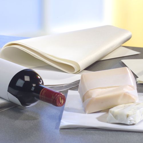 Une bouteille de vin rouge et des blocs de fromage sont partiellement enveloppés dans PAPSTAR 10 kg de papiers d'emballage, cellulose blanche de la PAPSTAR GmbH, en arrière-plan on peut voir une pile de feuilles de papier ciré sur la table.