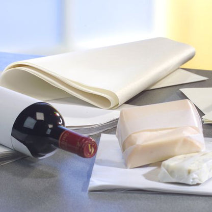 Une bouteille de vin rouge et des blocs de fromage sont partiellement enveloppés dans PAPSTAR 10 kg de papiers d'emballage, cellulose blanche de la PAPSTAR GmbH, en arrière-plan on peut voir une pile de feuilles de papier ciré sur la table.