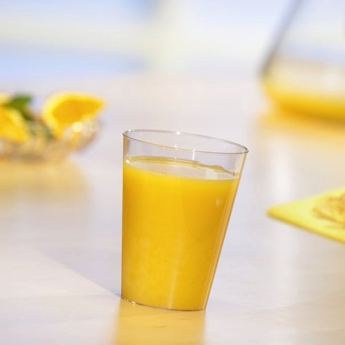 Un gobelet Starpak 10, PS 0,2l clair comme du cristal, rempli de jus d'orange se tient sur une table claire. En arrière-plan, des tranches d'orange et une cruche sont visibles de manière floue.