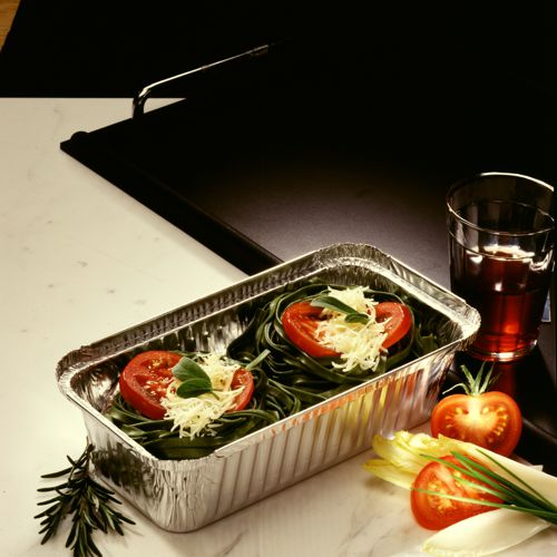 Un plateau Starpak 25, Alu + couvercle intérieur, revêtu de PP contient des nids de pâtes vertes non cuites avec du fromage, des tranches de tomate et du basilic. À côté : un verre de boisson rouge, des tomates, des endives et du romarin. Idéal pour le catering ou à emporter.
