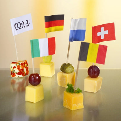 Six cubes de fromage sont garnis de raisins ou d'herbes et décorés pour une présentation élégante et écologique avec les petits drapeaux de décoration blancs de 8 cm de PAPSTAR GmbH, portant les inscriptions « Mild », Allemagne, Italie, Finlande, Suisse ou Belgique sur chaque drapeau.