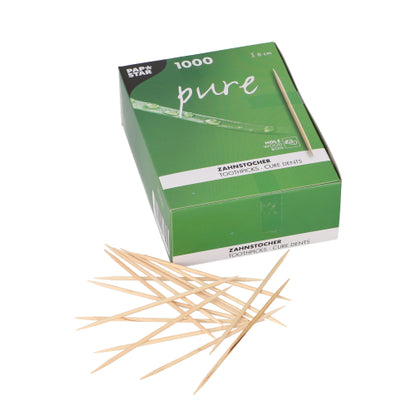 Une boîte PAPSTAR verte et blanche avec l'inscription "pure" contient 1000 cure-dents ronds en bois (8 cm), dont certains sont éparpillés à l'avant. Produit : PAPSTAR Cure-dents "pure" en bois, ronds, 8 cm | Boîte pliante (1000 pièces). Marque : PAPSTAR GmbH.