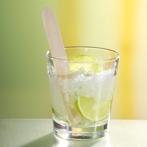 Un verre avec de la glace pilée, des tranches de citron vert et un PAPSTAR agitateur à boissons en bois de 15 cm (idéal pour les cocktails Caipirinha) de la PAPSTAR GmbH est posé sur une surface réfléchissante devant un fond flou jaune et vert.