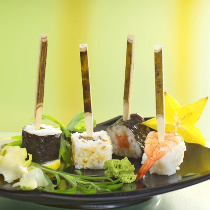 Sur un plateau en carton PAPSTAR (200 pièces), trois finger foods de sushi sont posés sur des brochettes PAPSTAR (11 cm bois naturel), accompagnés de carambole, wasabi, gingembre et verdure - parfait pour les fêtes.