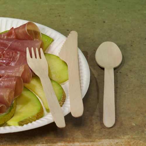 Une assiette en carton blanche avec des tranches de pomme verte et du prosciutto est posée sur une table. À côté se trouvent PAPSTAR cuillères en bois "pure" 15,7 cm - cuillères en bois écologiques et stables (100 pièces), de PAPSTAR GmbH, soigneusement disposées sur la surface marron.