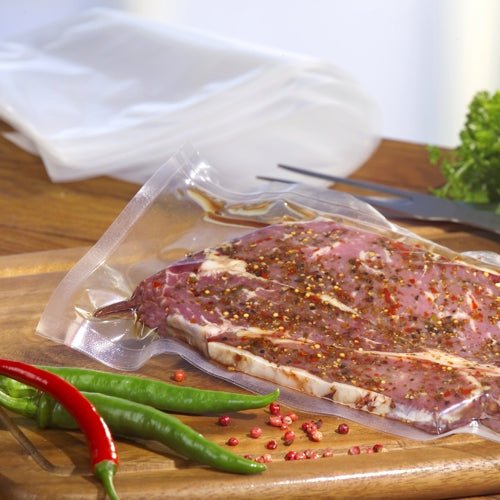 Un morceau de viande crue épicée est mis sous vide dans un sachet à souder Starpak 100 (PA/PE transparent) à l'aide d'une machine à vide à cloche. Il est posé sur une planche à découper en bois avec des piments rouges et verts et des légumes-feuilles à proximité.