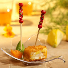 Un bol en verre transparent montre un cube alimentaire pané et deux morceaux de fruits, chacun avec un PAPSTAR Fingerfood Picker "Mexico" (7,5 cm) de PAPSTAR GmbH, parfait pour les fêtes. En arrière-plan légèrement flou, on peut voir des boissons et un quartier de citron.
