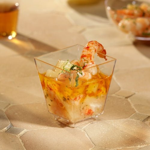Un bol à fingerfood Starpak 50 (PS transparent) de Starpak, rempli de crevettes et de cocktail de fruits de mer et garni d'herbes, est posé sur une surface texturée beige. D'autres tasses sont floues en arrière-plan - parfait pour des événements élégants.