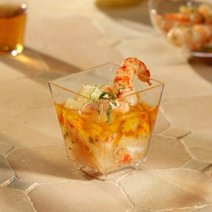 Un bol à fingerfood Starpak 50 (PS transparent) de Starpak, rempli de crevettes et de cocktail de fruits de mer et garni d'herbes, est posé sur une surface texturée beige. D'autres tasses sont floues en arrière-plan - parfait pour des événements élégants.