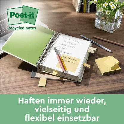 Ein Schreibtisch mit einem aufgeschlagenen Notizbuch, mehreren gelben Post-it® Super Sticky 100% Recycling Notes (47,6 x 47,6 mm) von 3M Deutschland GmbH, die auf den Seiten befestigt sind, einem Stift, einem Stapel dieser PEFC-zertifizierten Notizen (70 Blatt/Block) und einer Topfpflanze.