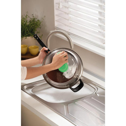 Quelqu'un nettoie un couvercle de casserole en métal au-dessus d'un évier savonneux avec l'éponge de nettoyage Scotch-Brite® Classic de 3M Deutschland GmbH, près d'une fenêtre avec des stores blancs et un comptoir avec une plante et des bols jaunes empilés.