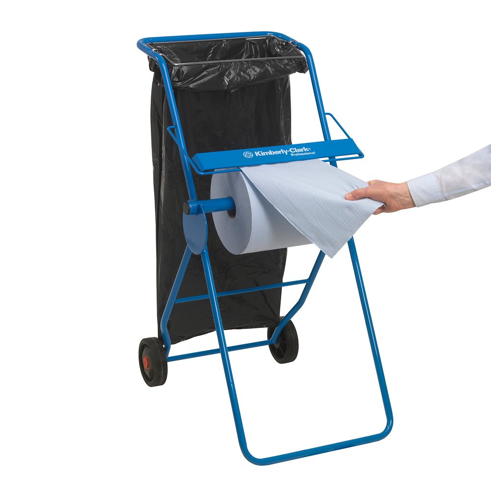 Un support métallique bleu avec deux roues tient un sac poubelle noir et un grand rouleau de lingettes WypAll® L10 EXTRA (1 rouleau x 1500 feuilles); une personne tire une feuille du rouleau. Sur la surface horizontale du support, l'inscription "Kimberly-Clark GmbH" est apposée.