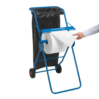 Un distributeur métallique bleu tient un papier essuie-tout WypAll® L10 pour surfaces - grand rouleau (38 x 23,5 cm) de Kimberly-Clark GmbH et un sac poubelle noir ; une personne tire une feuille pour le nettoyage des surfaces.