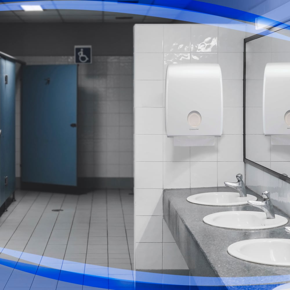 Une toilette publique avec trois lavabos, des distributeurs de serviettes pliées Kimberly-Clark Aquarius™, des distributeurs de savon hygiéniques, des miroirs sur le côté droit et des cabines bleues sur le côté gauche - une cabine ouverte est accessible aux fauteuils roulants.