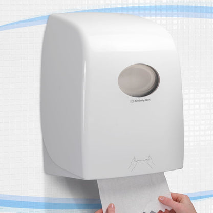 Le distributeur de serviettes en rouleau Aquarius™ - Blanc de Kimberly-Clark GmbH est monté sur le mur de carreaux, une main tirant une serviette par le bas. Il est idéal pour les espaces publics et dispose d'une fenêtre ronde en haut et d'instructions claires en dessous.