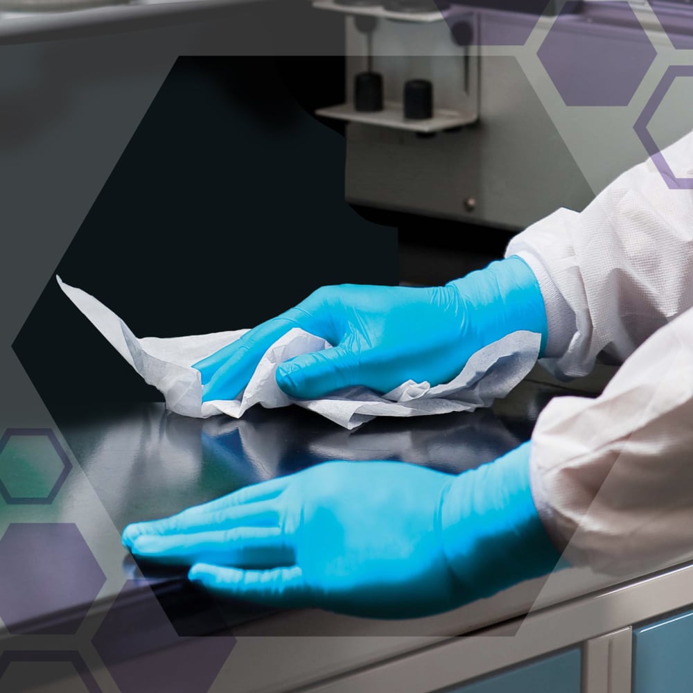 Eine Person mit blauen Handschuhen und einem weißen Laborkittel reinigt eine dunkle Labortischplatte mit Kimberly-Clark GmbH Auslaufartikel: Kimtech® Saugfähige Pflegetücher - Weiß (20 Säcke x 50 Blatt). Im Hintergrund sind Laborgeräte und sechseckige grafische Overlays zu sehen.