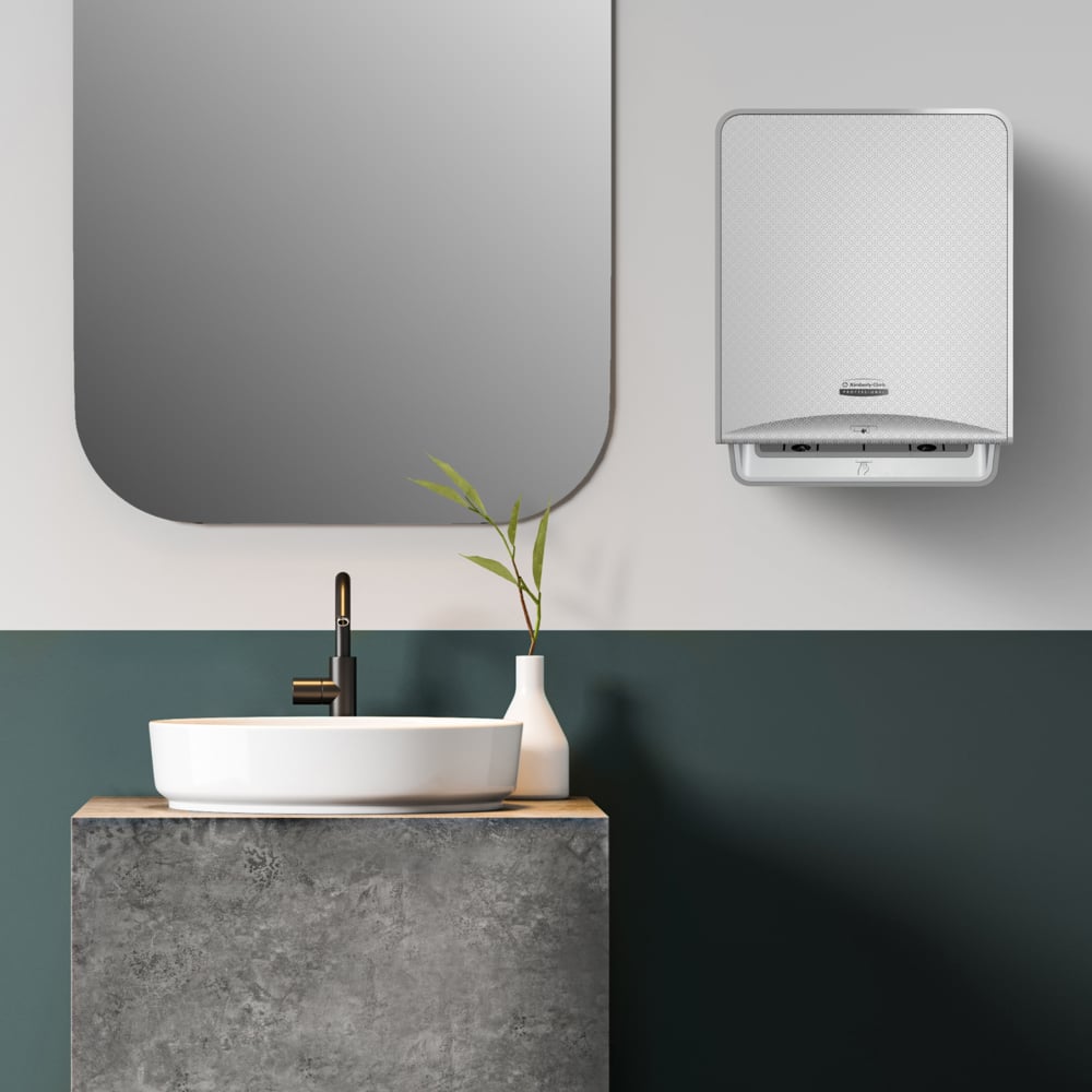 Salle de bain moderne avec miroir rectangulaire, lavabo blanc sur un meuble-lavabo en structure de béton, robinet noir, vase blanc avec branches vertes et un distributeur mural pour essuie-mains en rouleau Kleenex® de Kimberly-Clark GmbH.