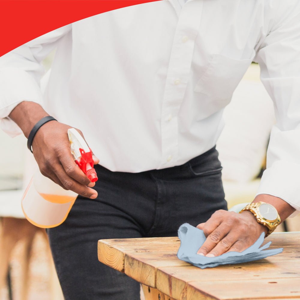 Une personne portant une chemise blanche et une montre nettoie une table en bois avec un chiffon bleu, peut-être WypAll® L20 General Clean - BRAG™ Box de Kimberly-Clark GmbH. L'image a une bordure rouge incurvée en haut qui souligne l'attention aux détails.