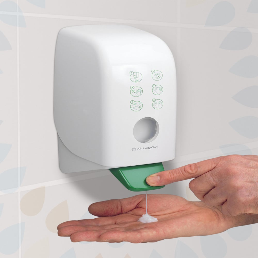 Une personne distribue de la lotion hydratante pour les mains et le corps Kleenex® (Kimberly-Clark GmbH) à partir d'une cartouche murale blanche de 1 litre dans une salle de bain carrelée avec un levier vert pour le soin des mains et du corps. Emballage : 6 cartouches par carton.