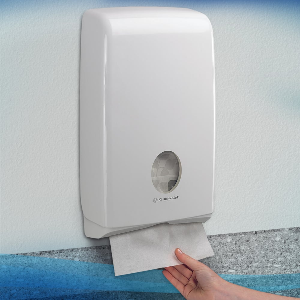 Eine Hand zieht ein Scott® SLIMFOLD Handtuch aus einem weißen Kimberly-Clark GmbH Wandspender mit Sichtfenster, das gefaltete Tücher im Inneren zeigt. Der hygienische Spender ist an einer hellen Wand montiert; er fasst 16 Clips x 110 Blatt pro Karton.