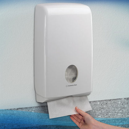 Eine Hand zieht ein Scott® SLIMFOLD Handtuch aus einem weißen Kimberly-Clark GmbH Wandspender mit Sichtfenster, das gefaltete Tücher im Inneren zeigt. Der hygienische Spender ist an einer hellen Wand montiert; er fasst 16 Clips x 110 Blatt pro Karton.