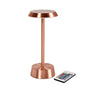 La lampe LED NOUR sans fil en or rose de Duni GmbH a une base ronde et un dessus plat pour un style moderne. À côté de la lampe se trouve une télécommande avec des boutons colorés.