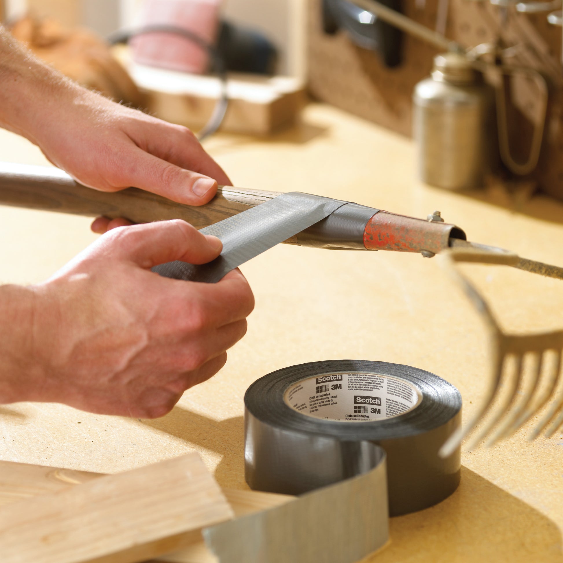 Eine Person repariert einen Stiel einer Gartenharke, indem sie ihn mit Scotch® Extremium™ All Weather Hochleistungs-Klebeband (27 m x 48 mm) der 3M Deutschland GmbH umwickelt. Eine offene Rolle und Werkzeug liegen auf der Werkbank daneben.