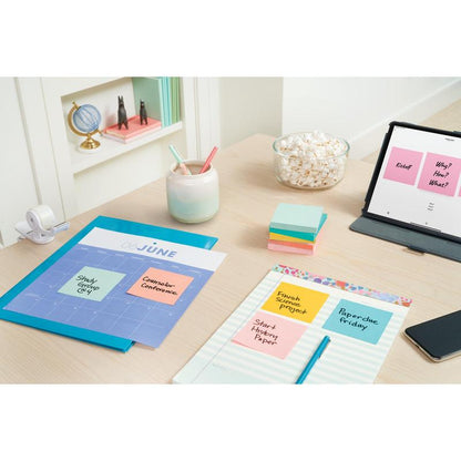 Un bureau avec un calendrier de juin, des stylos pastel dans un gobelet, des Notes Post-it® Beachside Collection (38x51mm, 100 feuilles/bloc, 12 blocs/paquet, emballage en carton, 100% PEFC), un dévidoir de ruban adhésif, un tableau ouvert avec des notes - et un petit globe sur l'étagère.