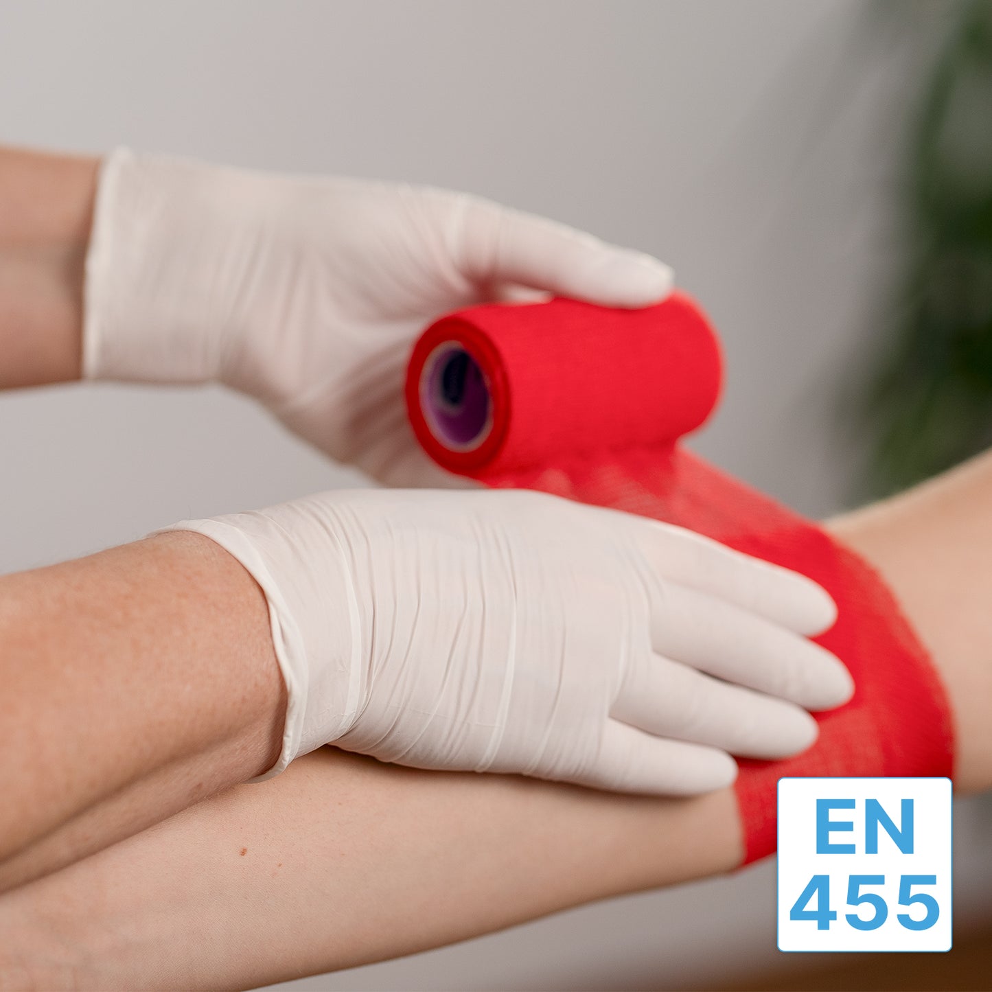 Une personne portant des gants en nitrile ARNOMED NITRIL WHITE 200er de ARNOWA GmbH enroule un bandage rouge autour du bras d'une autre personne. Dans le coin inférieur droit, une étiquette "EN 455" bleu-blanc est visible.