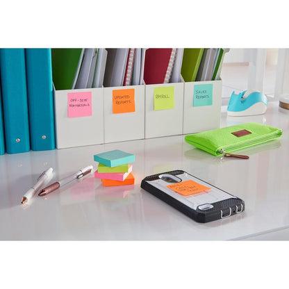 Sur le bureau bien rangé se trouvent des classeurs étiquetés, une trousse verte, deux stylos et un smartphone avec un Post-it® Super Sticky Note (76x127 mm, 3M Deutschland GmbH) sur lequel est écrit "Appel client Chet AM". La surface bien éclairée est blanche.