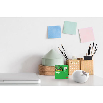 Un bureau avec un ordinateur portable fermé, un distributeur de ruban de table Scotch™ Karim Rashid de 3M Deutschland GmbH et une boîte de ruban adhésif, des contenants pour stylos et crayons, un petit ornement en forme de maison et trois notes autocollantes colorées sur le mur blanc.