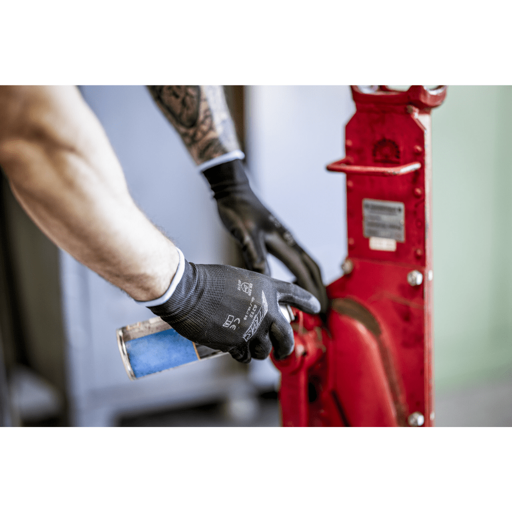 Une personne portant des gants en nylon NITRAS noirs, enduits de PU par AS Arbeitsschutz GmbH, nettoie ou lubrifie avec une bombe de pulvérisation bleue une pièce de machine en métal rouge. Seuls les bras et les mains sont visibles.
