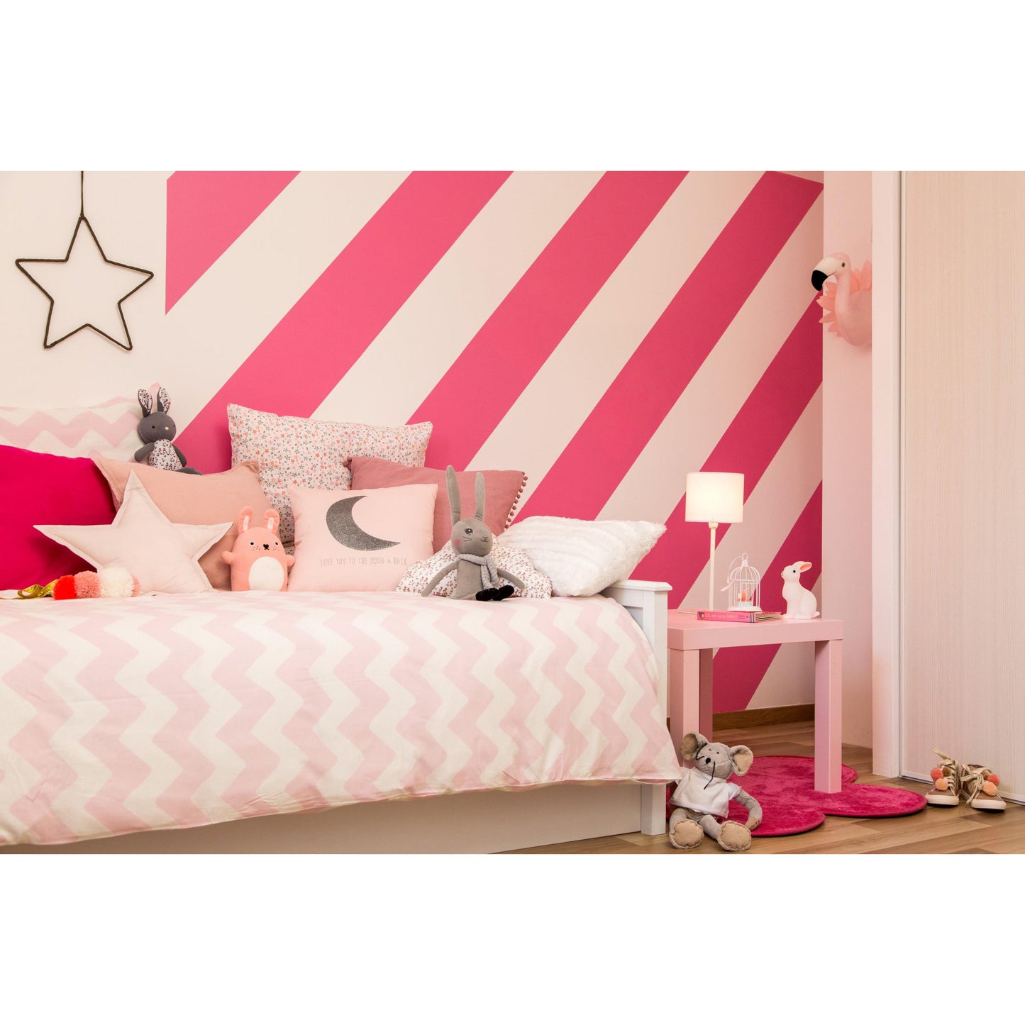 Ein Kinderzimmer mit weißem Bett, rosa und grauen Kissen, Plüschtieren und kräftigen rosa-weiß diagonal gestreiften Wänden, gestrichen mit ScotchBlue™ Malerabdeckband für scharfe Linien von 3M Deutschland GmbH. Eine Lampe, ein Tisch, Spielzeug und Schuhe vervollständigen den gemütlichen Raum.