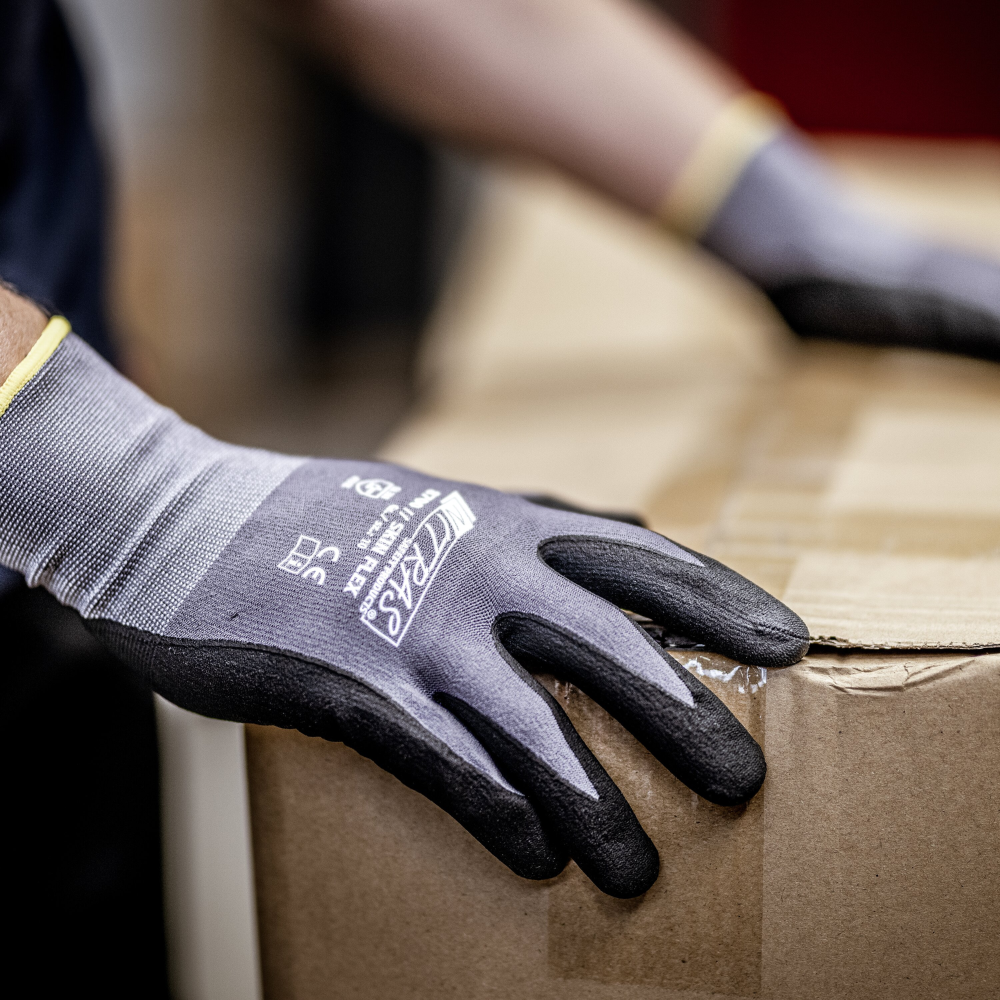 Une personne porte des gants tricotés NITRAS SKIN FLEX de AS Arbeitsschutz GmbH en tricot spécial stretch, gris, partiellement enduits, soulève un carton scellé. Seules les mains gantées et une partie du carton sont visibles.