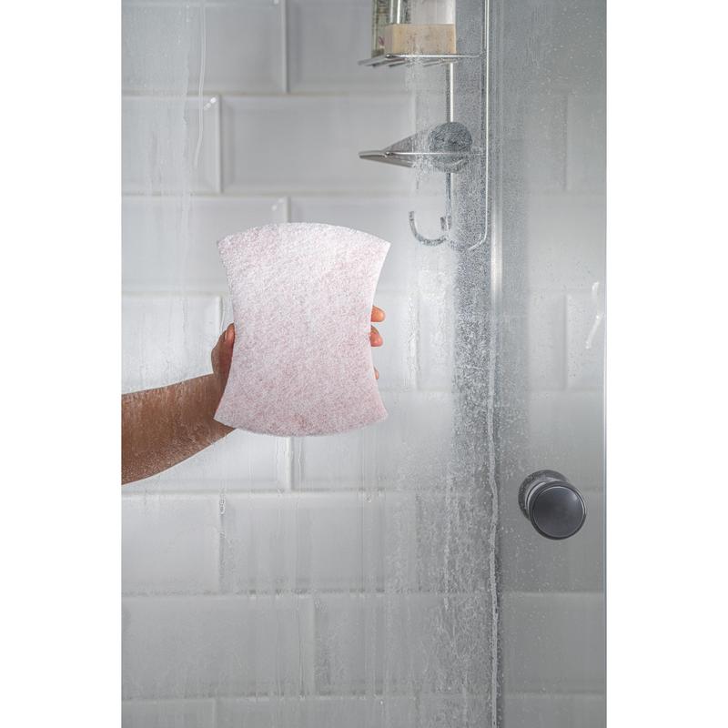 Une main utilise l'éponge à poignée Scotch-Brite® Bath XXL de 3M Deutschland GmbH dans une douche en verre avec des carreaux métro blancs et une pomme de douche en métal, car les traces d'eau sur la porte soulignent la nécessité d'un nettoyage efficace de la salle de bain.