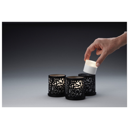 Une main place une petite bougie cylindrique dans un des trois bougeoirs Twine 75 x Ø 75 mm Noir de Duni GmbH, ornés de découpes élaborées, idéaux pour un design moderne. La décoration de table est placée devant un arrière-plan avec un dégradé de couleurs allant du gris foncé au gris clair.