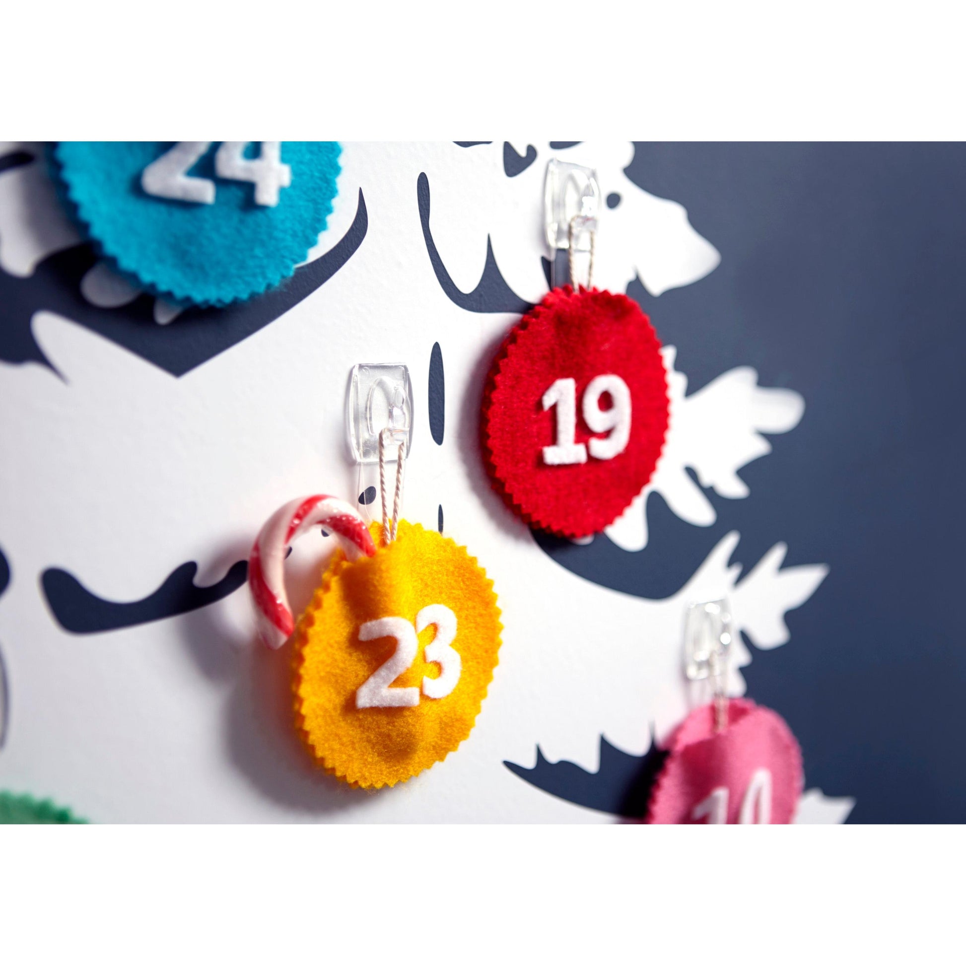 Nahaufnahme einer weißen Tafel mit Filzkreisen mit den Nummern 19, 23 und 24, die jeweils an transparenten Command™ Dekorations-Clips der 3M Deutschland GmbH befestigt sind; an dem Clip der Nummer 23 hängt eine kleine Zuckerstange.