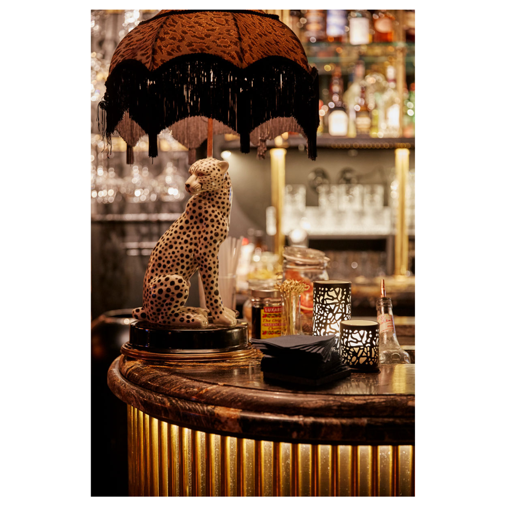 Un bar faiblement éclairé avec une statue de léopard sur le comptoir sous un abat-jour à franges se distingue par un design moderne. Des bouteilles et des serviettes entourent un élégant bougeoir Twine 75 x Ø 75 mm Noir de Duni GmbH, à côté d'une lanterne en métal, créant une décoration de table accueillante.
.
