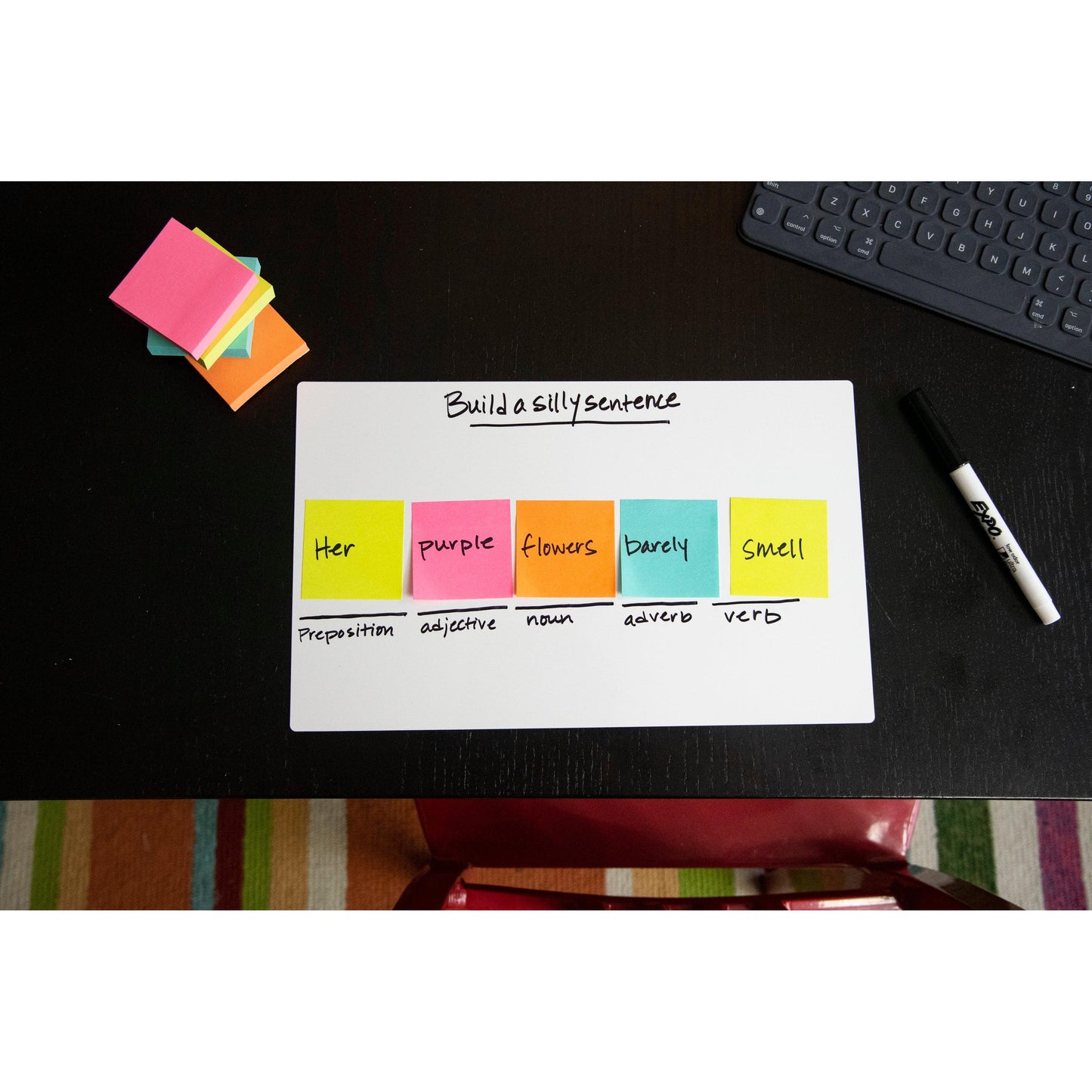 Auf einem schwarzen Schreibtisch befindet sich ein Whiteboard mit einer bunten Tabelle zum Thema "Bilde einen dummen Satz" mit Wörtern auf 3M Post-it® Super Sticky Z-Notes in Neonfarben (76x127 mm, 100 Blatt/Block, 6 Blöcke/Packung), daneben ein Marker und zusätzliche Haftnotizen.