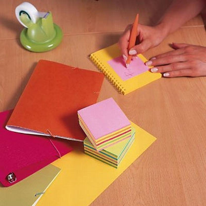 Quelqu'un écrit sur un cube de notes Post-it® rose de 3M Deutschland GmbH, Ultra-Couleurs, sur un bureau en bois avec des piles de notes adhésives de 76x76 mm (450 feuilles/bloc), un bloc-notes, des classeurs et un dévidoir de ruban adhésif vert.