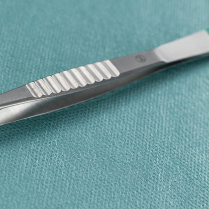 Un gros plan d'une pince à éclats Hartmann de Paul Hartmann AG droite de 9cm, stérile (paquet de 40 pièces), posée à côté d'un scalpel chirurgical en métal sur un tissu médical texturé couleur sarcelle.